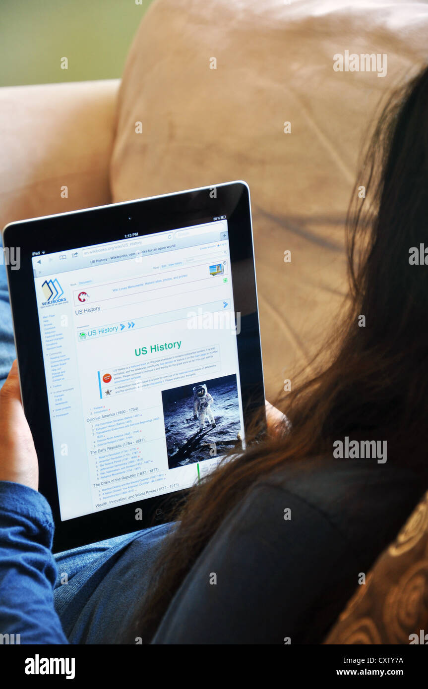 Young girl with iPad sitting on sofa at home. Wikibooks US History website shown on the iPad screen. Stock Photo