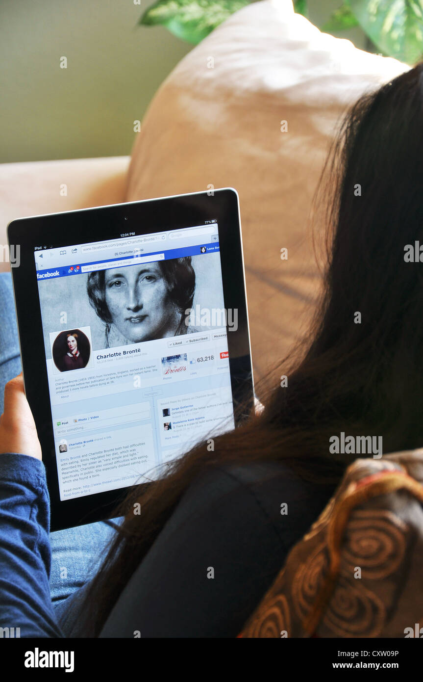 Young girl with iPad sitting on sofa at home. Charlotte Brontë fan club on Facebook shown on the iPad screen. Stock Photo