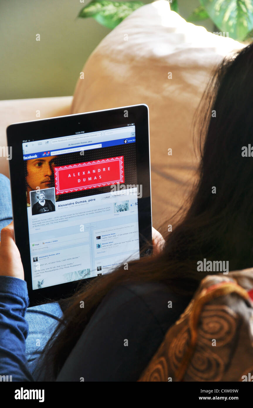 Young girl with iPad sitting on sofa at home. Alexandre Dumas fan club on Facebook shown on the iPad screen. Stock Photo