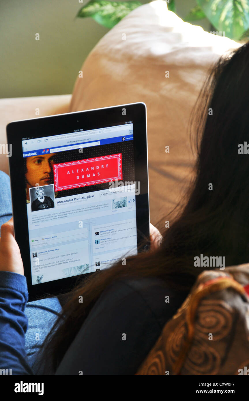 Young girl with iPad sitting on sofa at home. Facebook Alexandre Dumas fan website shown on the iPad screen. Stock Photo
