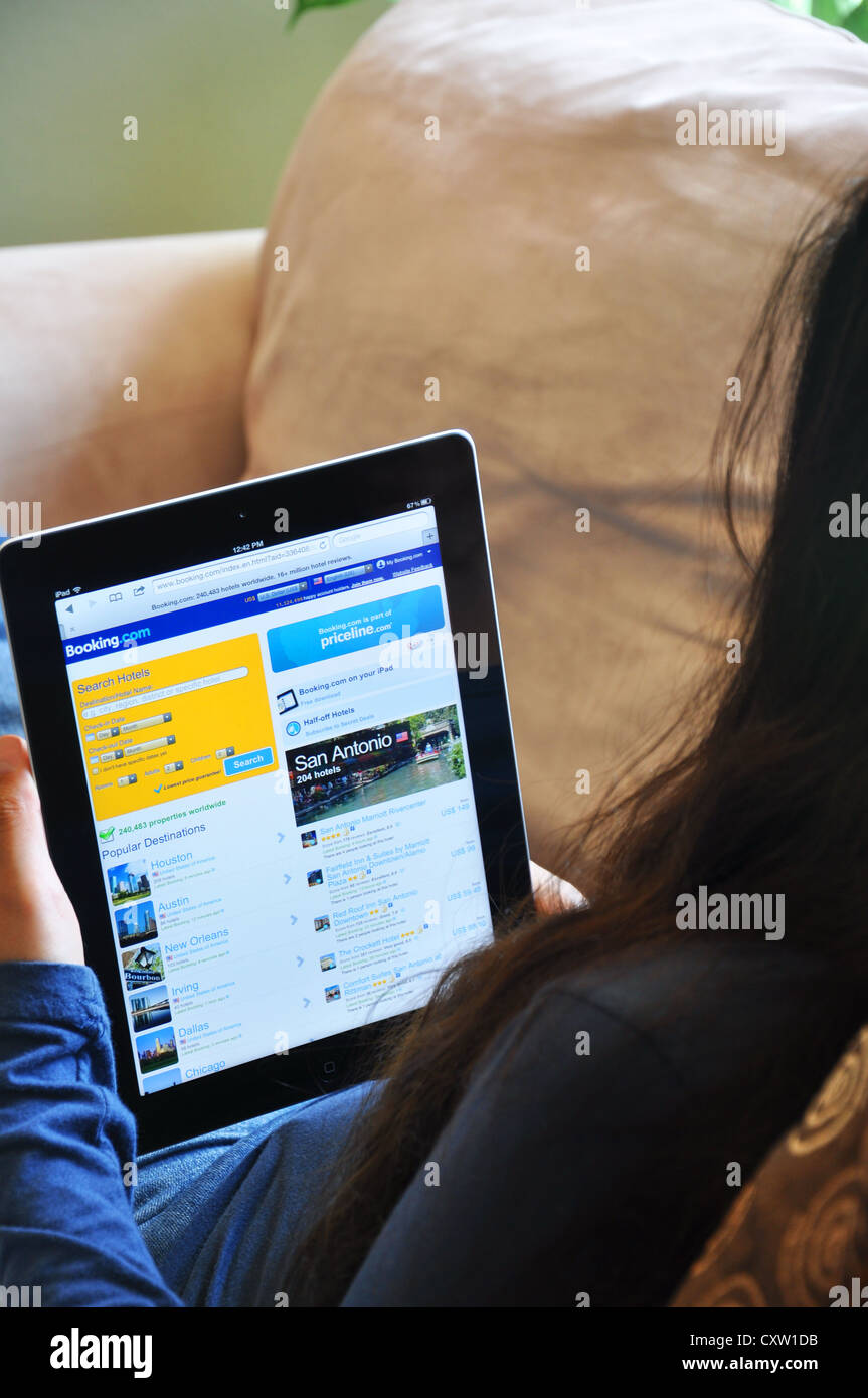 Young girl with iPad sitting on sofa at home. Booking.com hotel reservations website shown on the iPad screen. Stock Photo