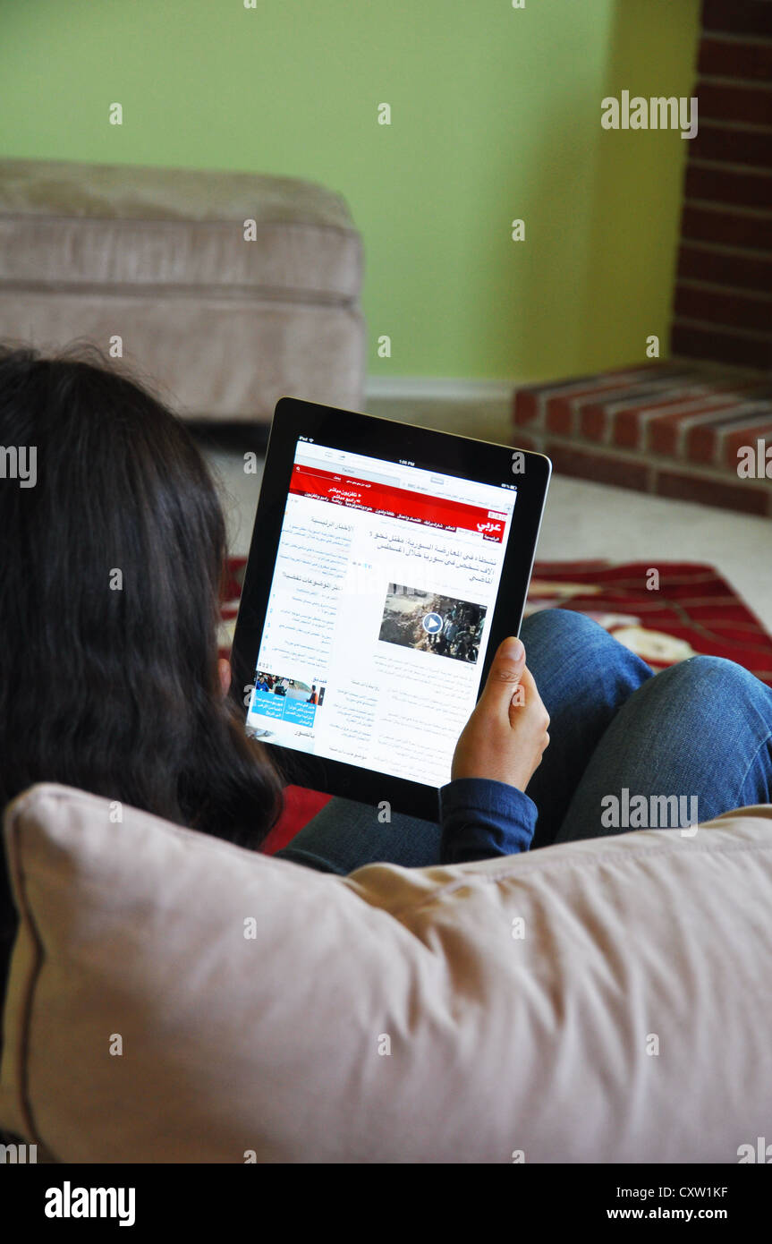 Young girl with iPad sitting on sofa at home. BBC in Arabic website shown on the iPad screen. Stock Photo