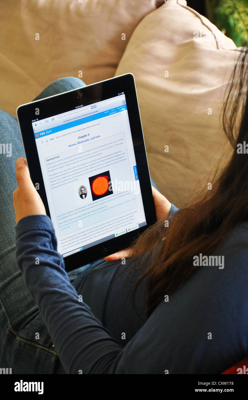 Female student  with iPad sitting on sofa at home. Science textbook shown on the iPad screen. Stock Photo