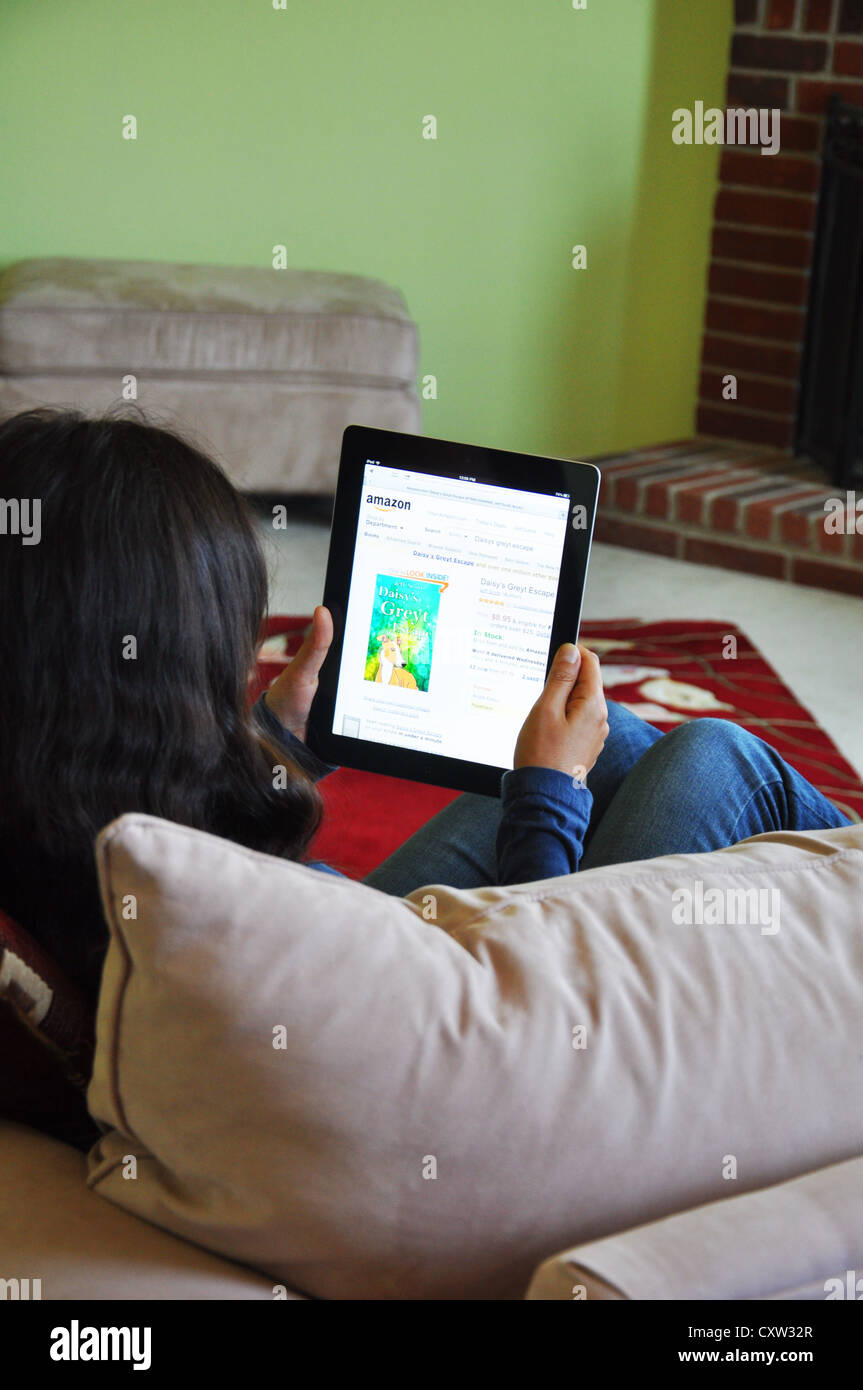 Young girl with iPad sitting on sofa at home. Amazon website shown on the iPad screen. Stock Photo