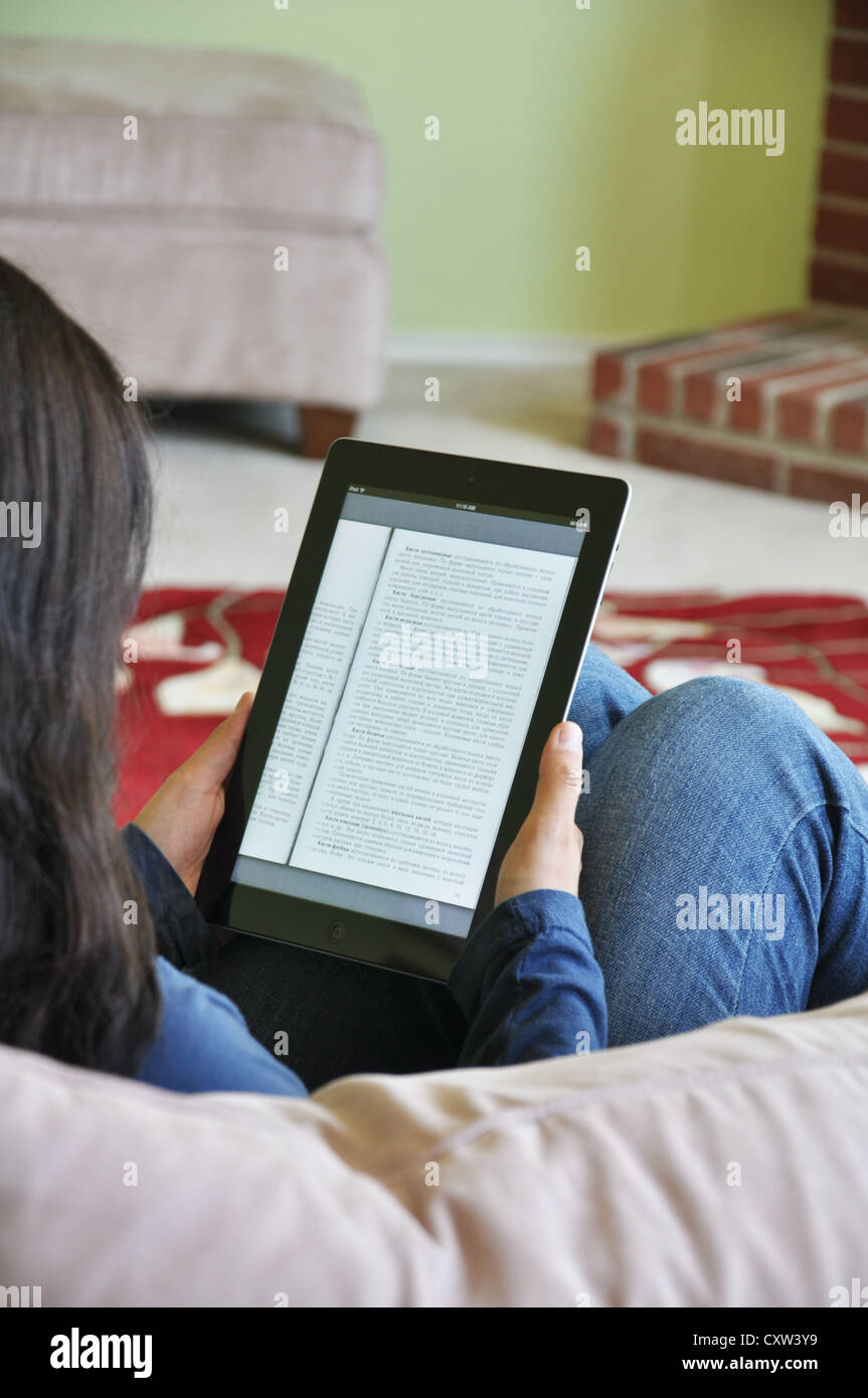 Young girl with iPad sitting on sofa at home. Student with Russian textbook shown on the iPad screen. Stock Photo