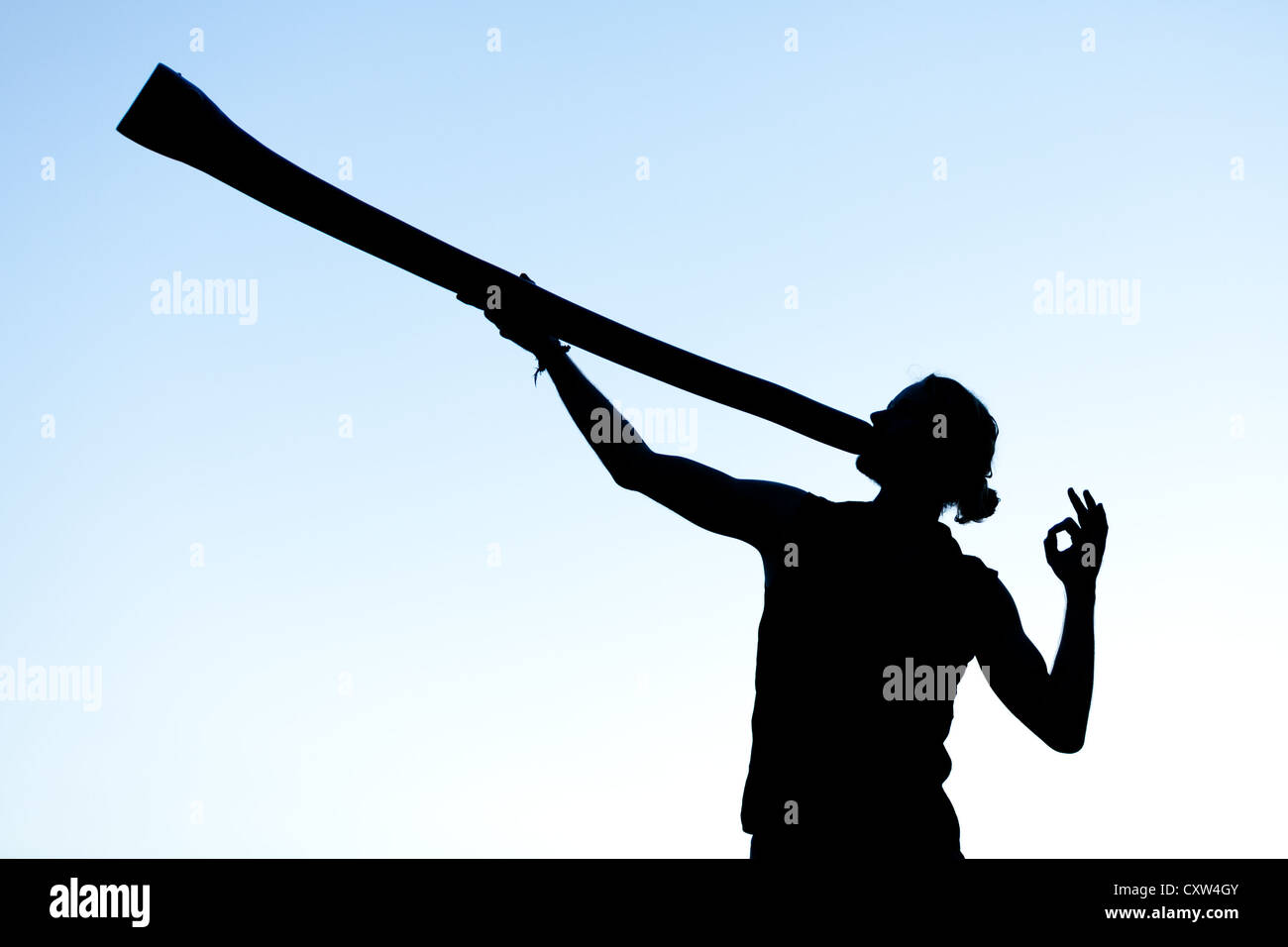 A didgeridoo player plays his didge while silhouetted against the evening sky. Hand forms a sacred mudra. Stock Photo