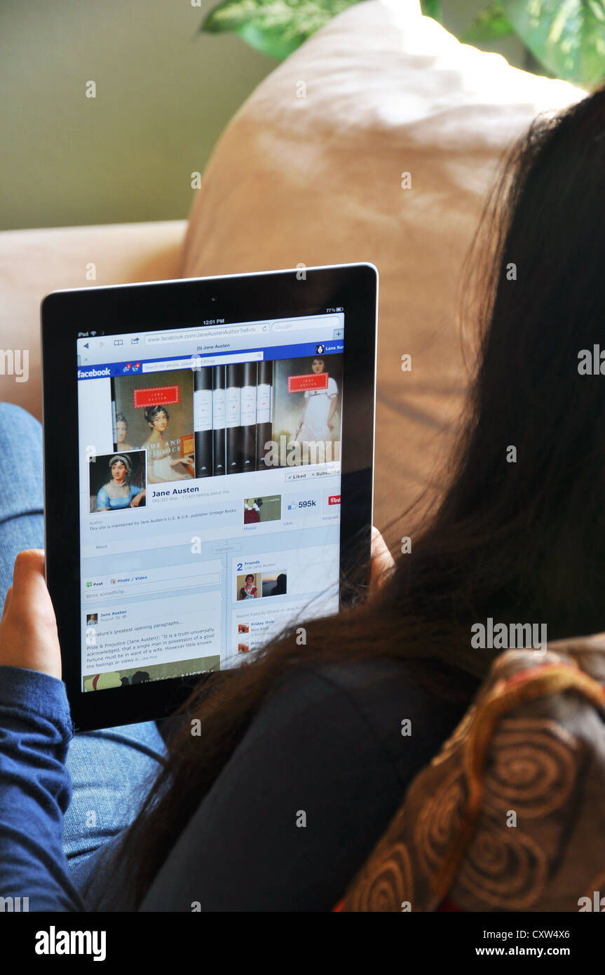 Young girl with iPad sitting on sofa at home. Facebook Jane Austen fan website shown on the iPad screen. Stock Photo