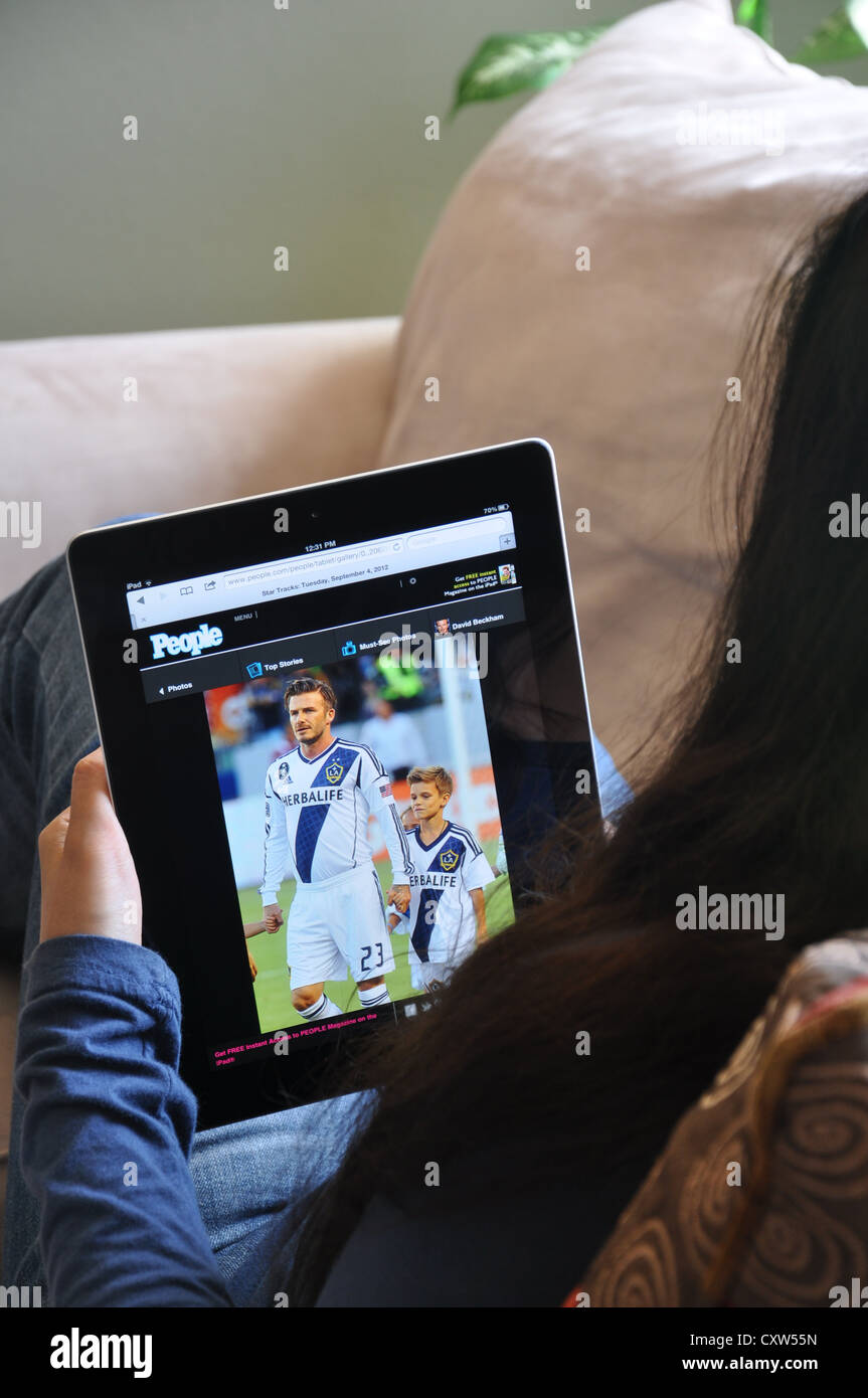 Young girl with iPad sitting on sofa at home. People magazine website shown on the iPad screen. Stock Photo