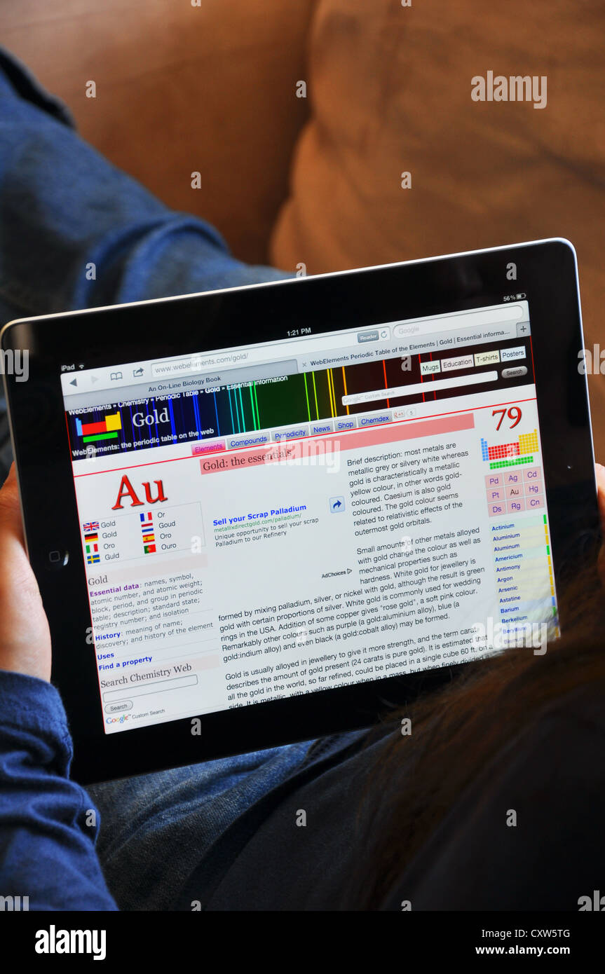 Female student with iPad sitting on sofa at home. Chemical elements - gold - online article shown on the iPad screen. Stock Photo