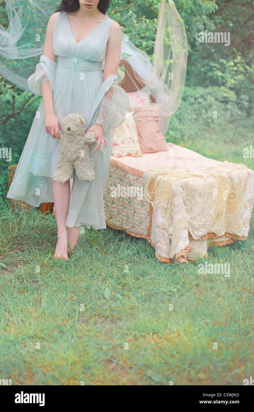 Cropped shot of a pale young woman wearing a fancy blue nightgown in the woods and holding a teddybear beside a child's bed Stock Photo
