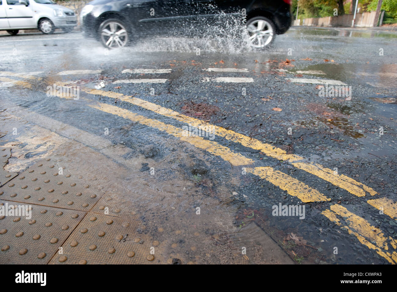 Burst water main pipe flooding road with cars going past Stock Photo