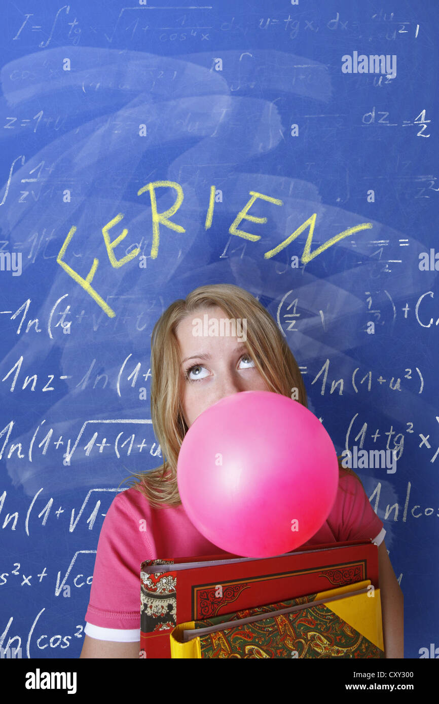 Schoolgirl blowing a bubble with bubble gum in front of a school blackboard with the word Ferien, German for holidays Stock Photo