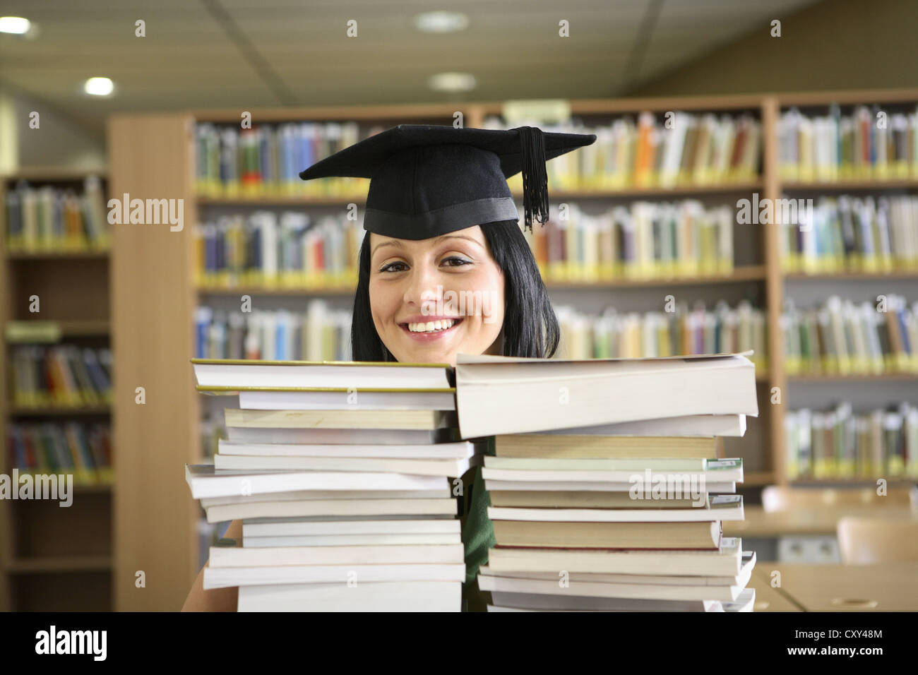 Smiling female student wearing a graduation cap behind stacks of books in a university library Stock Photo