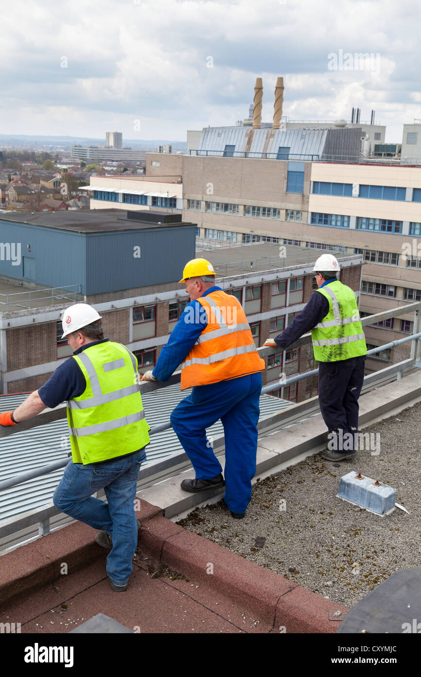 three workman in high visibility jackets and hard hats leaning against rail on roof waiting Stock Photo