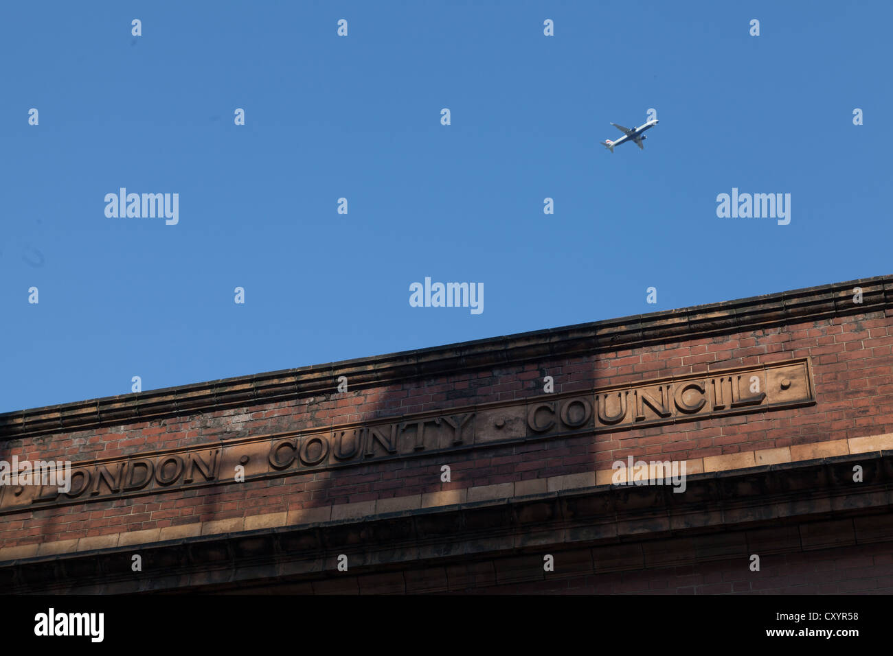 Plane flying through blue sky above old building with London County Council sign in brickwork Bermondsey, London Stock Photo