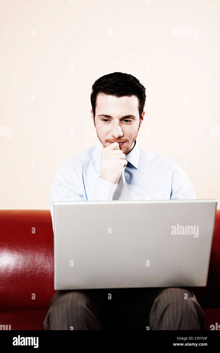 Young man sitting with his laptop on a sofa, thoughtful Stock Photo