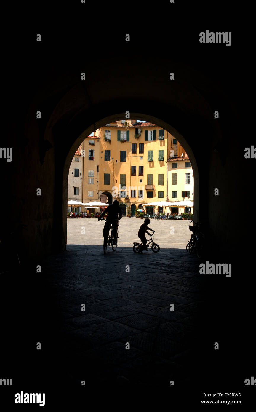 Adult and child cycling in shadow on a piazza in Italy shot through a archway Stock Photo