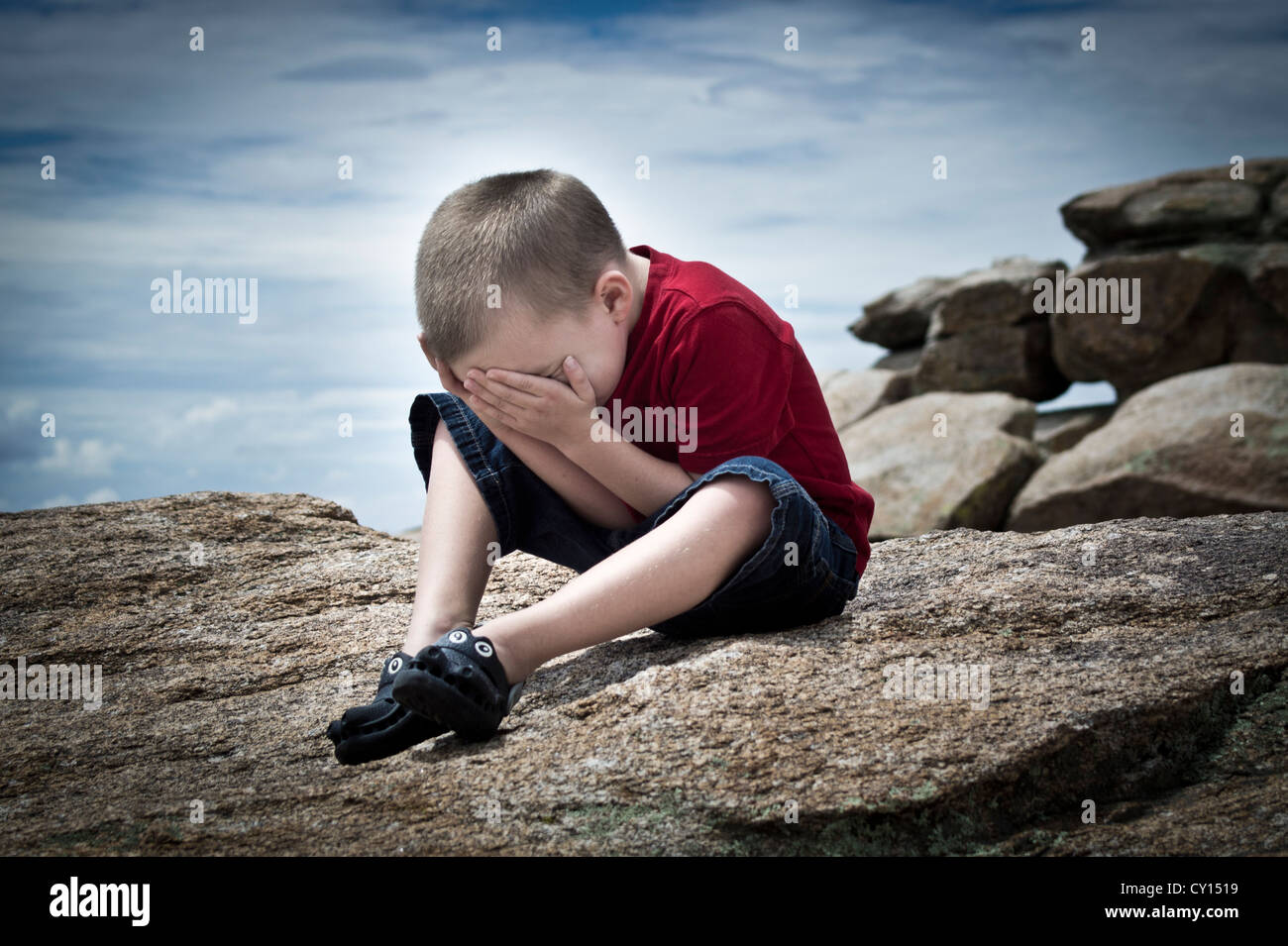 A five-year-old boy with autism feeling overwhelmed by his surroundings hides his face in his hands. Stock Photo