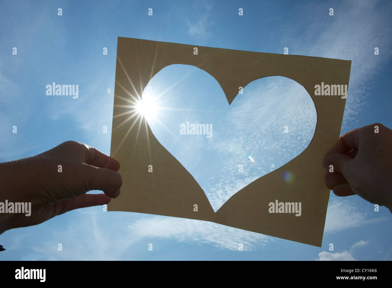 ein Herz aus Pappe in den Himmel gehalten gegen die Sonne, a heart cut of a piece of paper, two hands holding it against the sun Stock Photo
