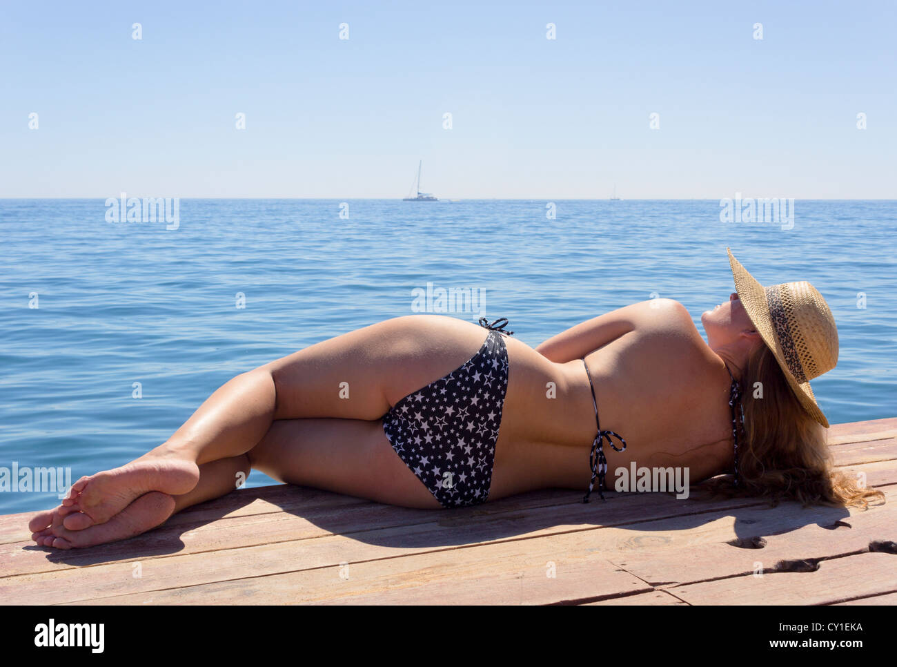 Young woman in black and white bikini and straw sunhat lying on wooden boardwalk. Stock Photo