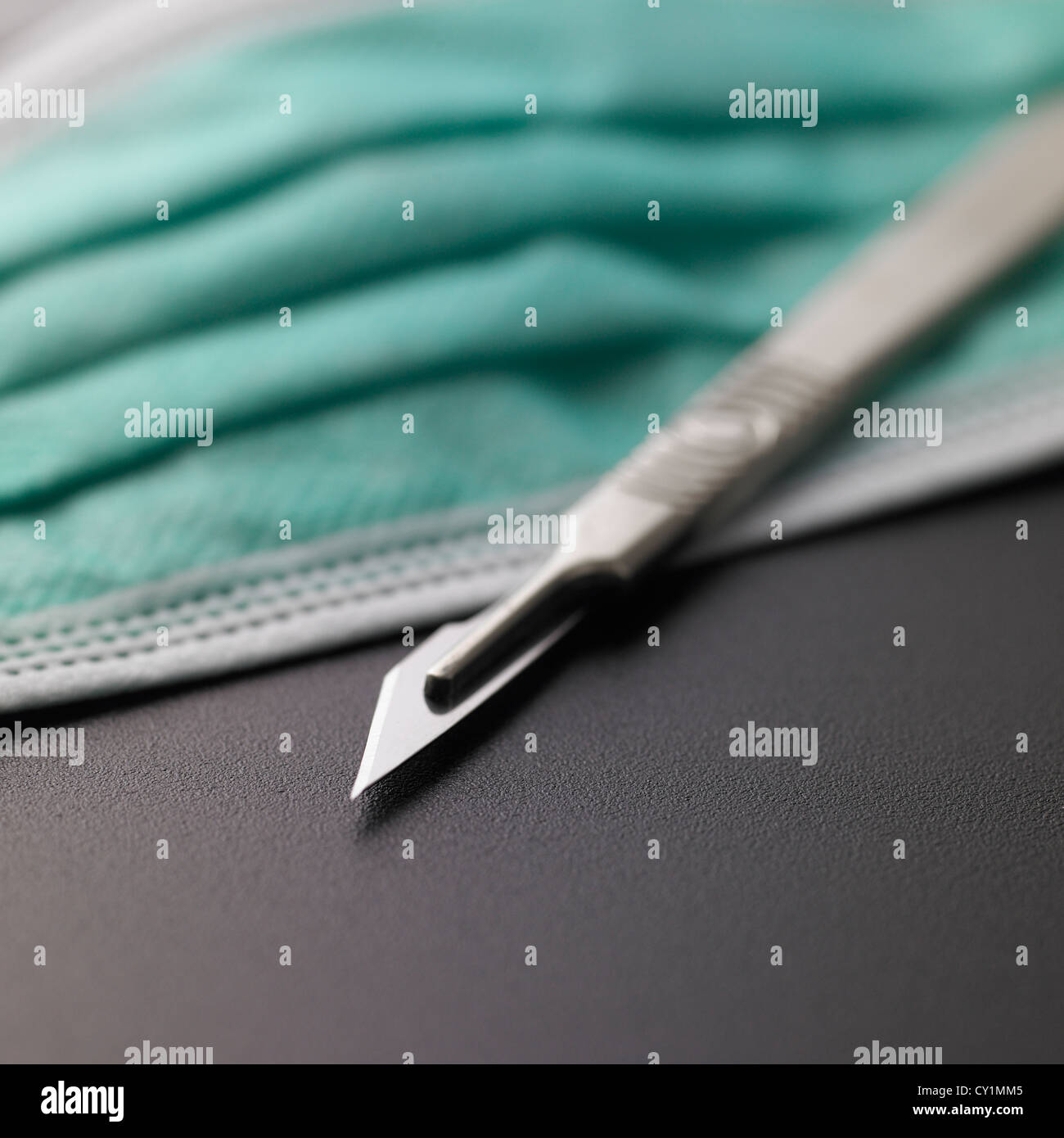 Surgical scalpel closeup above protective medical face mask on metallic grey background Stock Photo