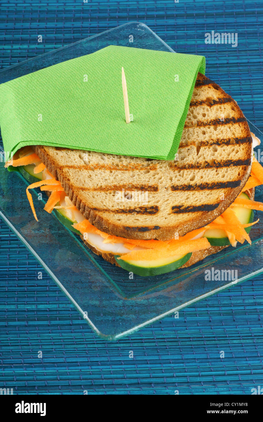 Roasted turkey sandwich with julienne carrots and slices of cucumber on a transparent glass plate. Selective focus. Stock Photo