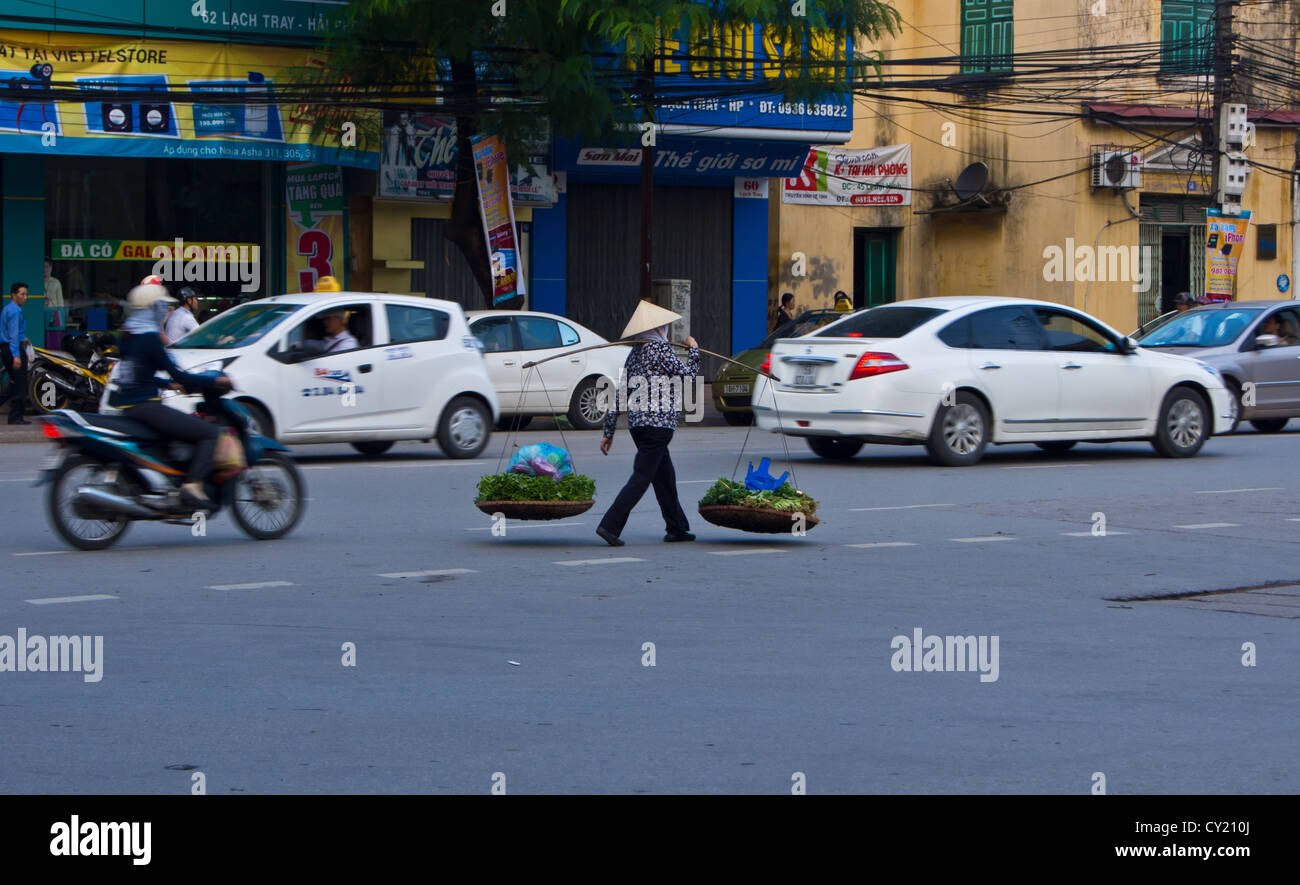 A Vietnamese lady walking along a busy road with her baskets of vegetables for sale  in Hai Phong Vietnam Stock Photo