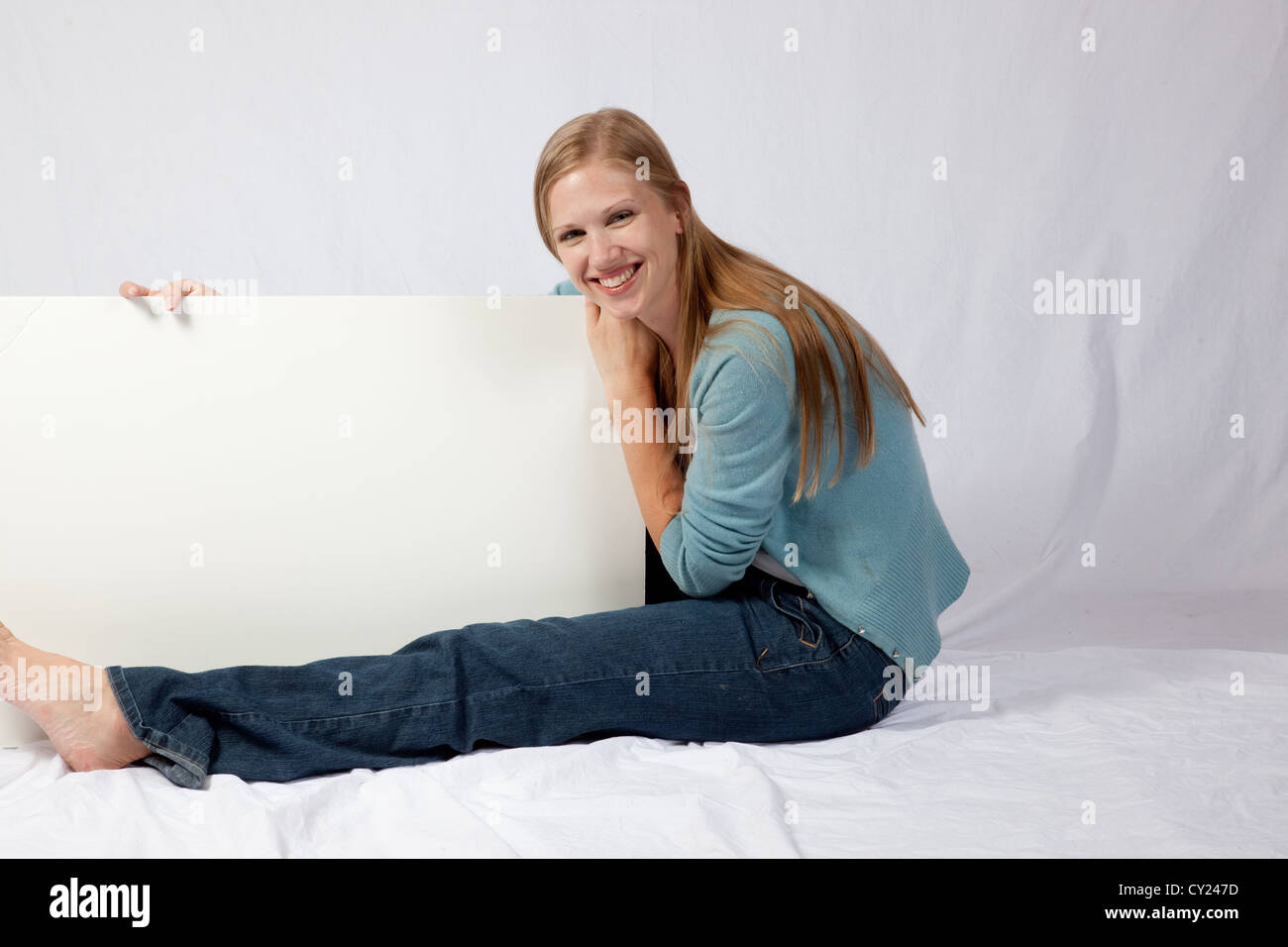 Pretty blond woman sitting and holding a white sign that is blank, ready for you to write your message on. Stock Photo