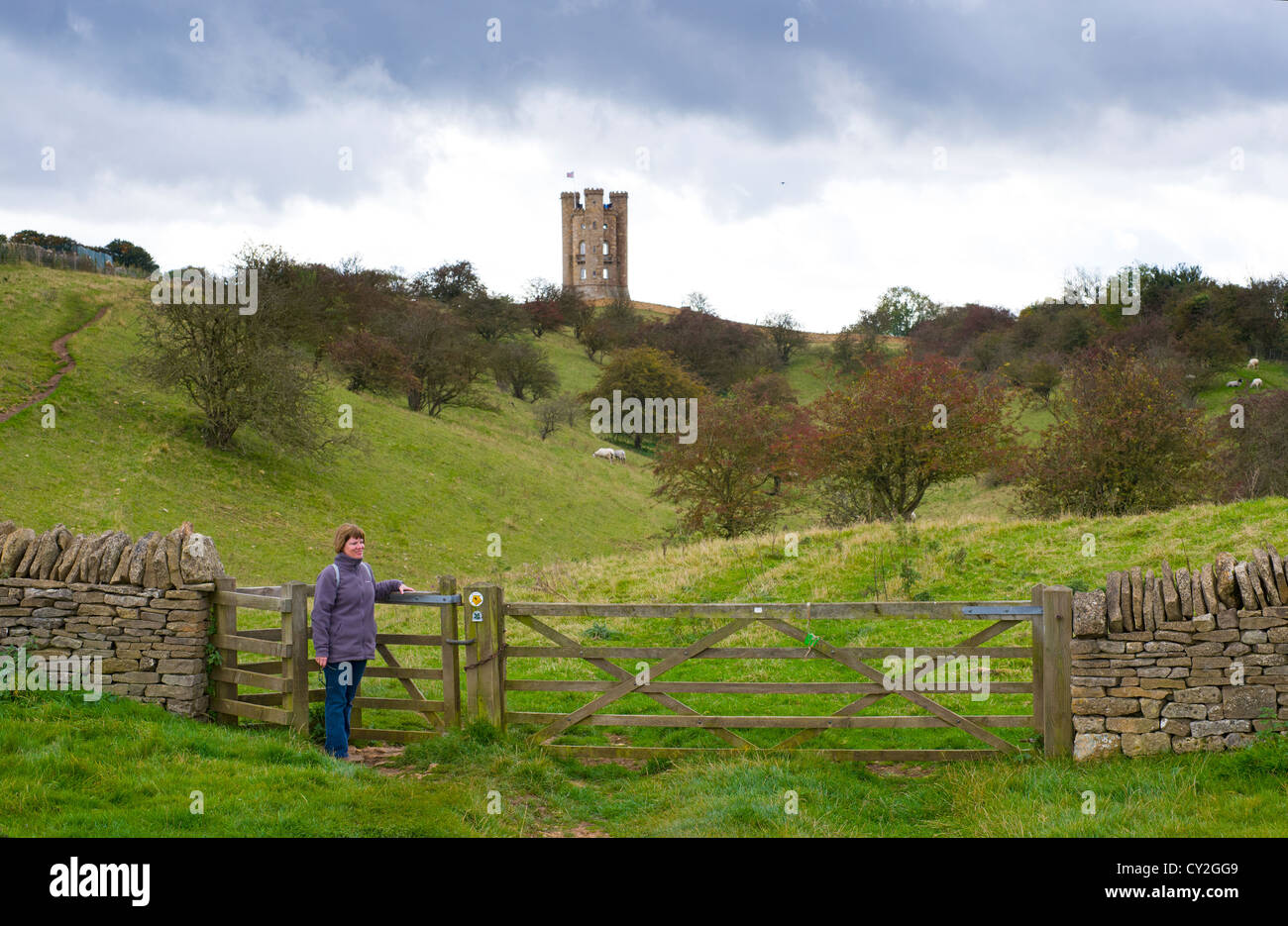 Female walker, at kissing gate on the Cotswold way footpath, with Broadway Tower folly on Broadway Hill in distance, Stock Photo