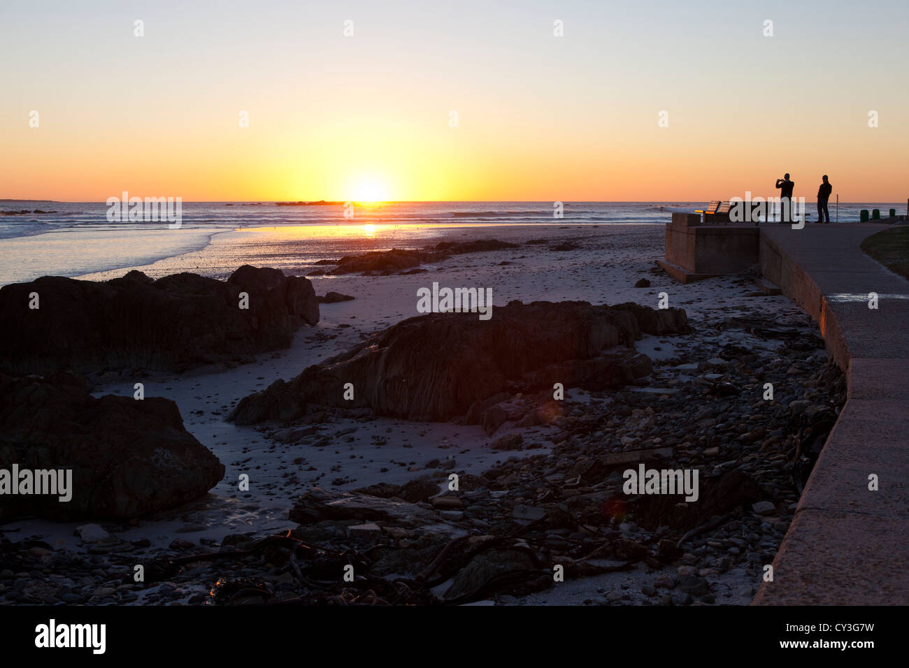 Two silhouettes of people watching the sunset and taking photos at Bloubergstrand. Stock Photo