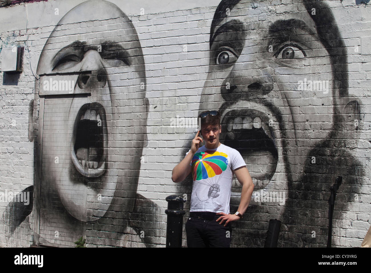 A fashionably dressed young man makes a call on his mobile telephone beneath a piece of graffiti depicting extremists Stock Photo