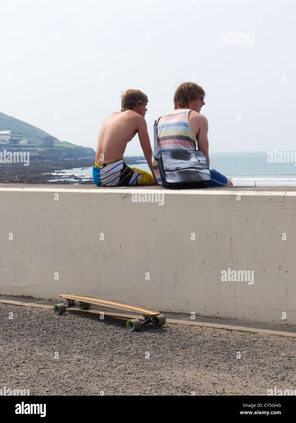 Young men with longboard skate board sit on wall overlooking beach. Westward Ho Devon. Stock Photo