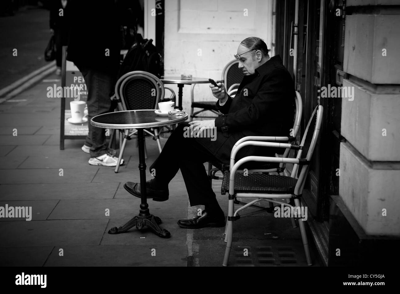 Man in black mac sitting outside cafe texting on phone Stock Photo