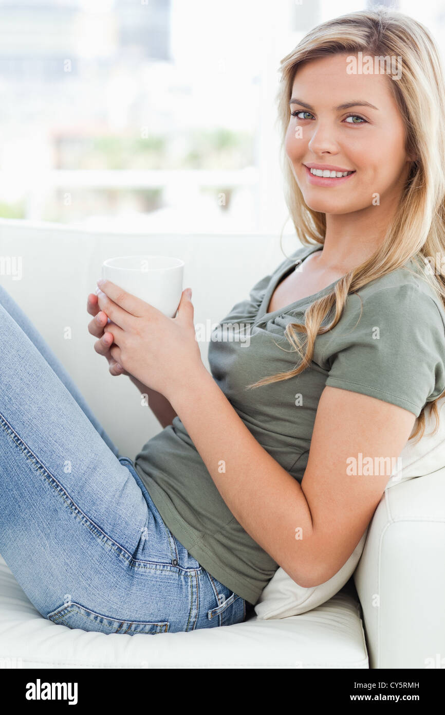 Close up, woman sitting across couch, cup in hands looking forward and smiling Stock Photo