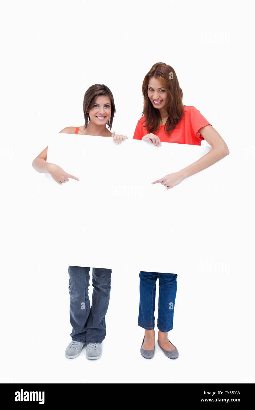 Smiling teenage girls standing upright behind a blank poster while pointing it Stock Photo