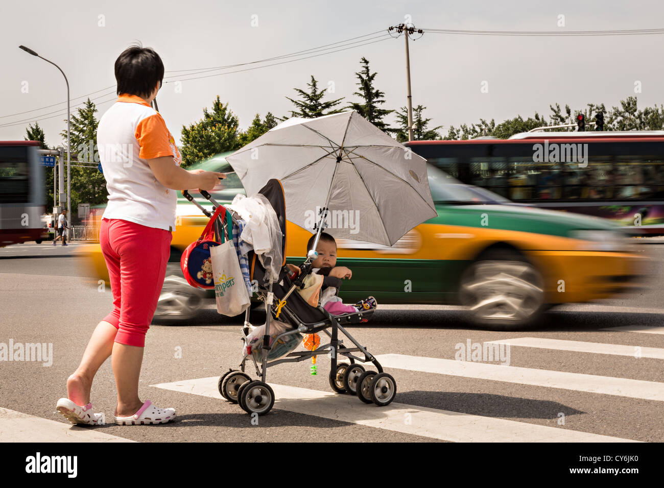 A Chinese woman pushing a baby carriage waits for traffic to cross a busy road during summer in Beijing, China Stock Photo