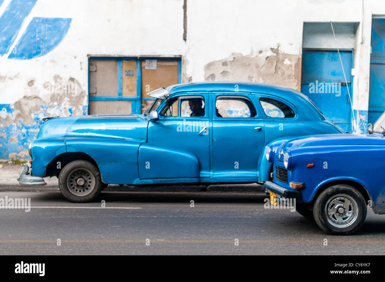 Two blue vintage American cars being driven in Havana with blue and white wall in the background. Stock Photo
