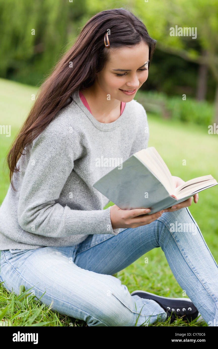 Young relaxed girl sitting on the grass in a park while reading a book ...