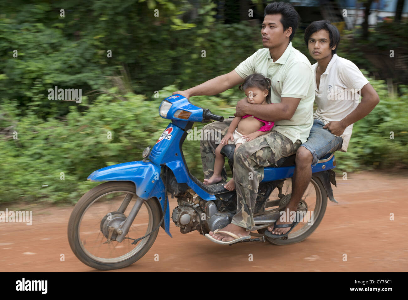 A father holds on to his sleeping child as he passes by on a motorbike in O'Ambel village, Sisophon, Cambodia Stock Photo