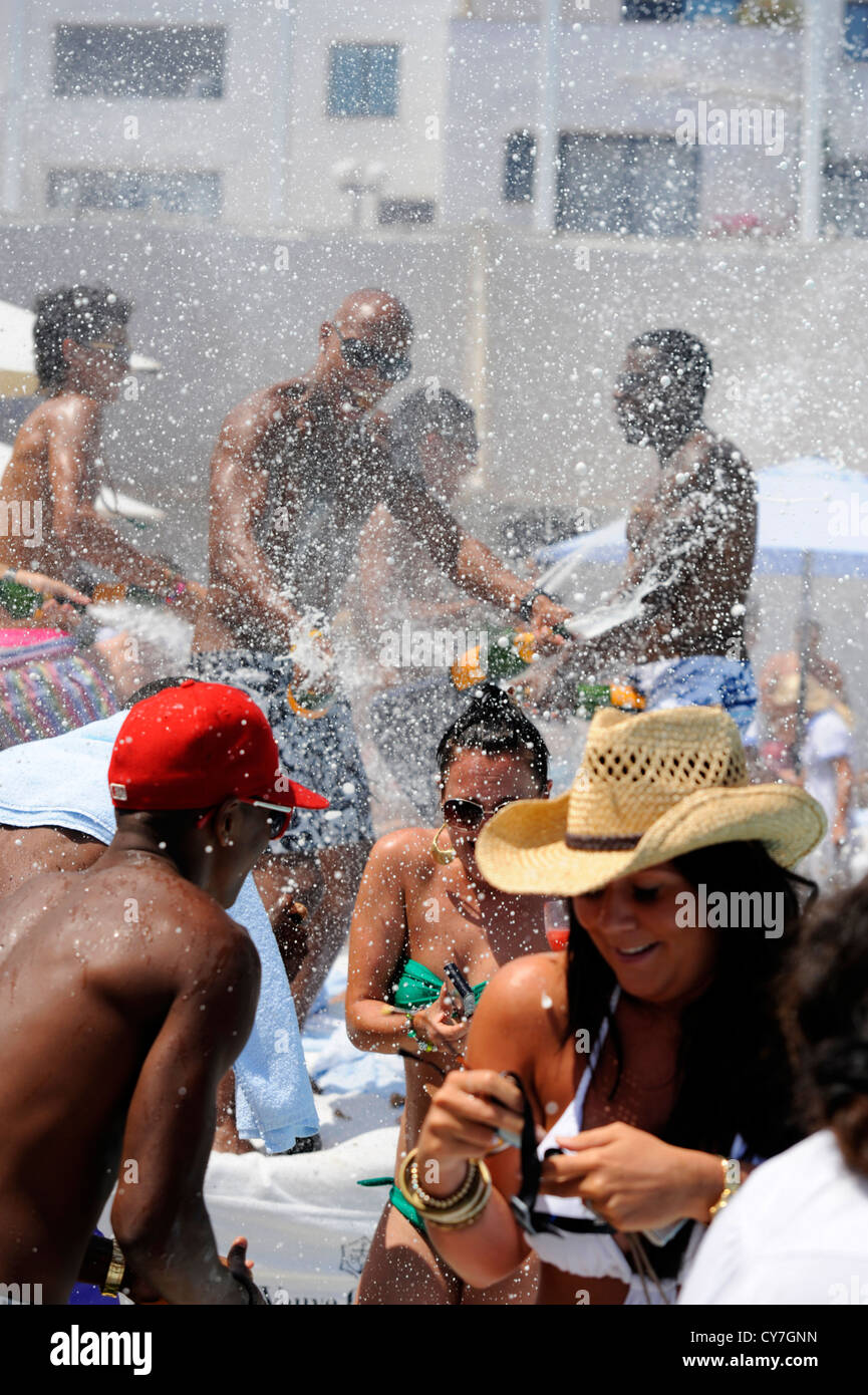 Party revelers standing on sun loungers at beach club spraying champagne during summer pool party Stock Photo