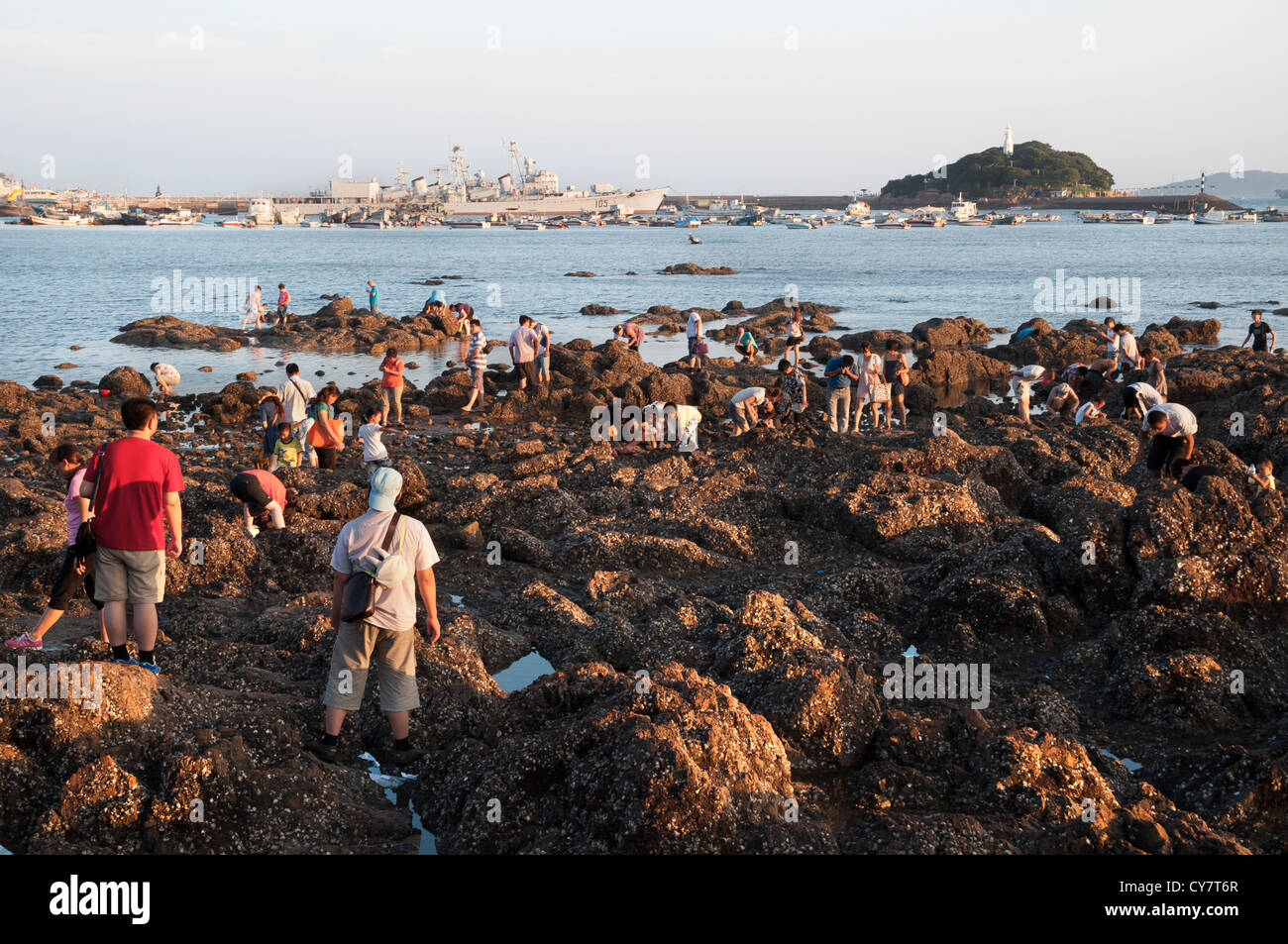 Holidaymakers search the rocks pools in Qingdao while a retired destroyer from the Qingdao Naval Museum sits in the background Stock Photo