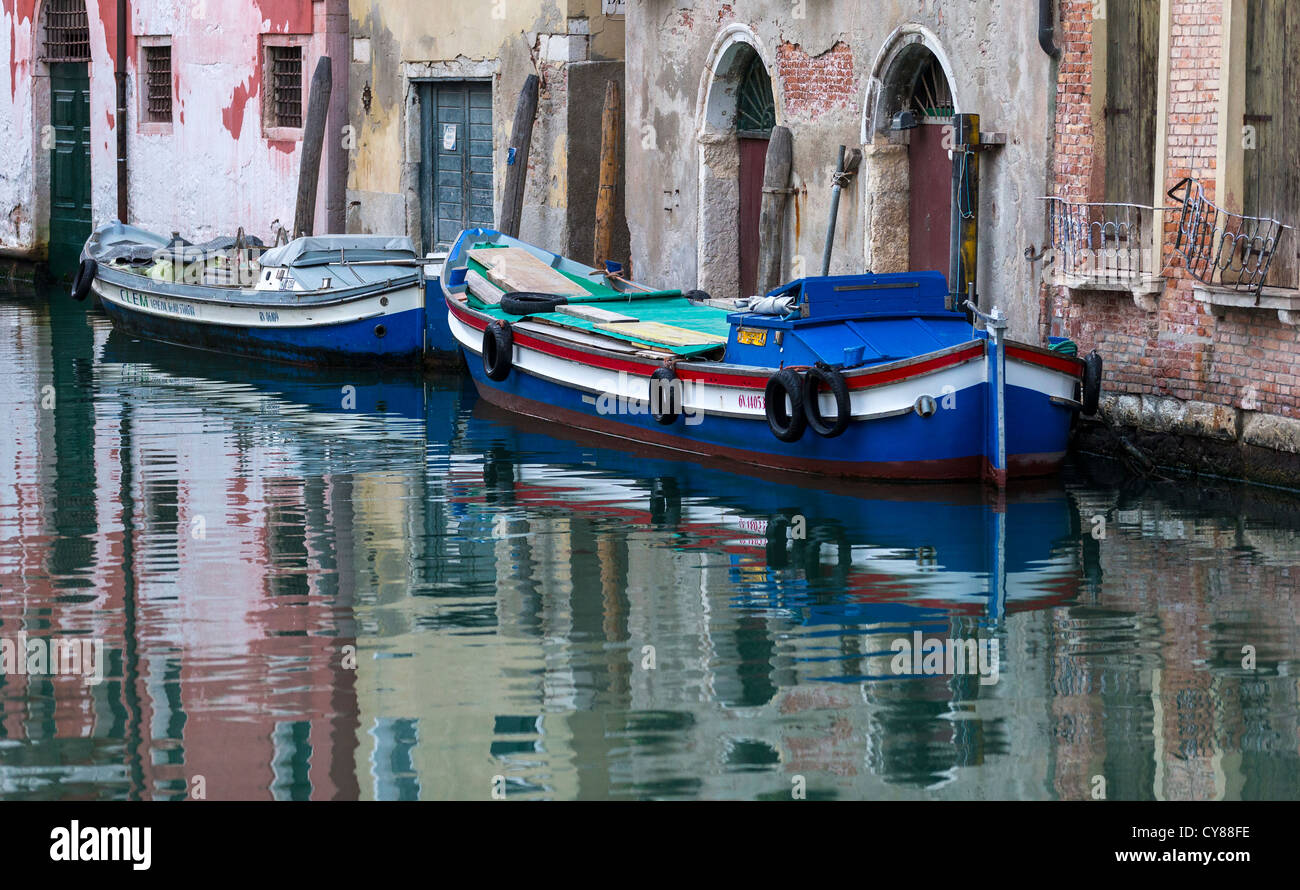 Faded pastel colours of dilapidated buildings contrast with brightly-painted boats, reflected in Venetian canal waters. Stock Photo
