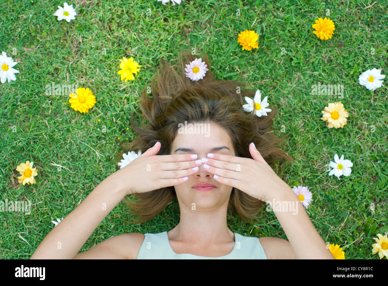 Woman lying on grass surrounded by flowers with hands covering eyes