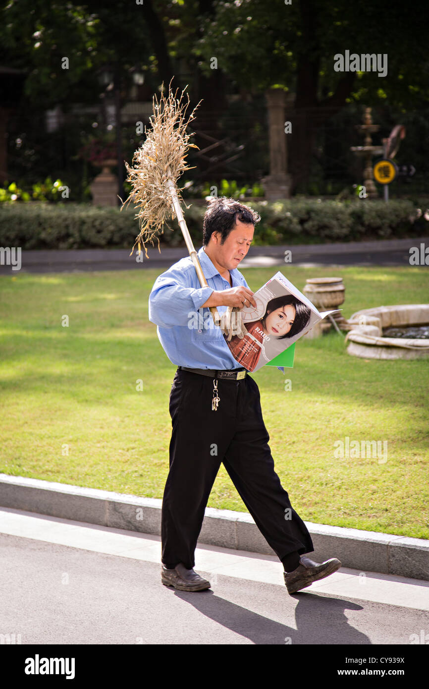 A Chinese workman carrying a broom reads a modern glossy magazine as he walks in Shanghai, China Stock Photo