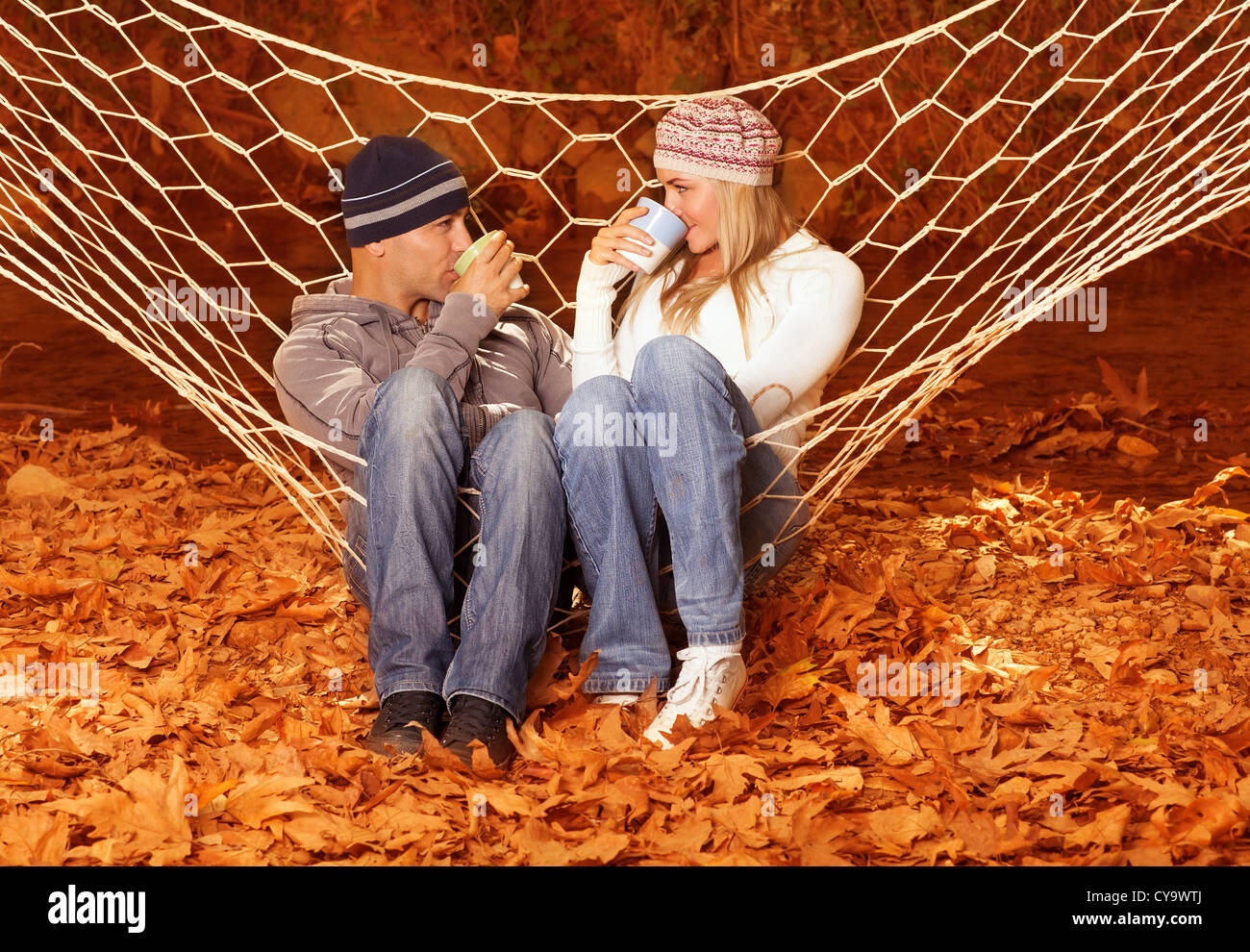 Image of beautiful young couple sitting in hammock and drink hot chokolate, lovely woman with handsome man relaxed outdoors Stock Photo