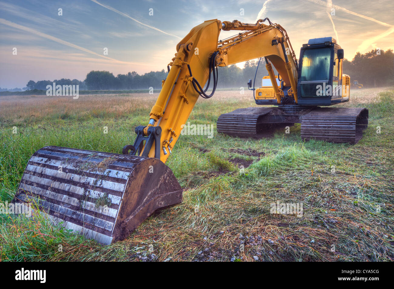 Yellow excavator in early morning light under a blue sky with condensation trails Stock Photo