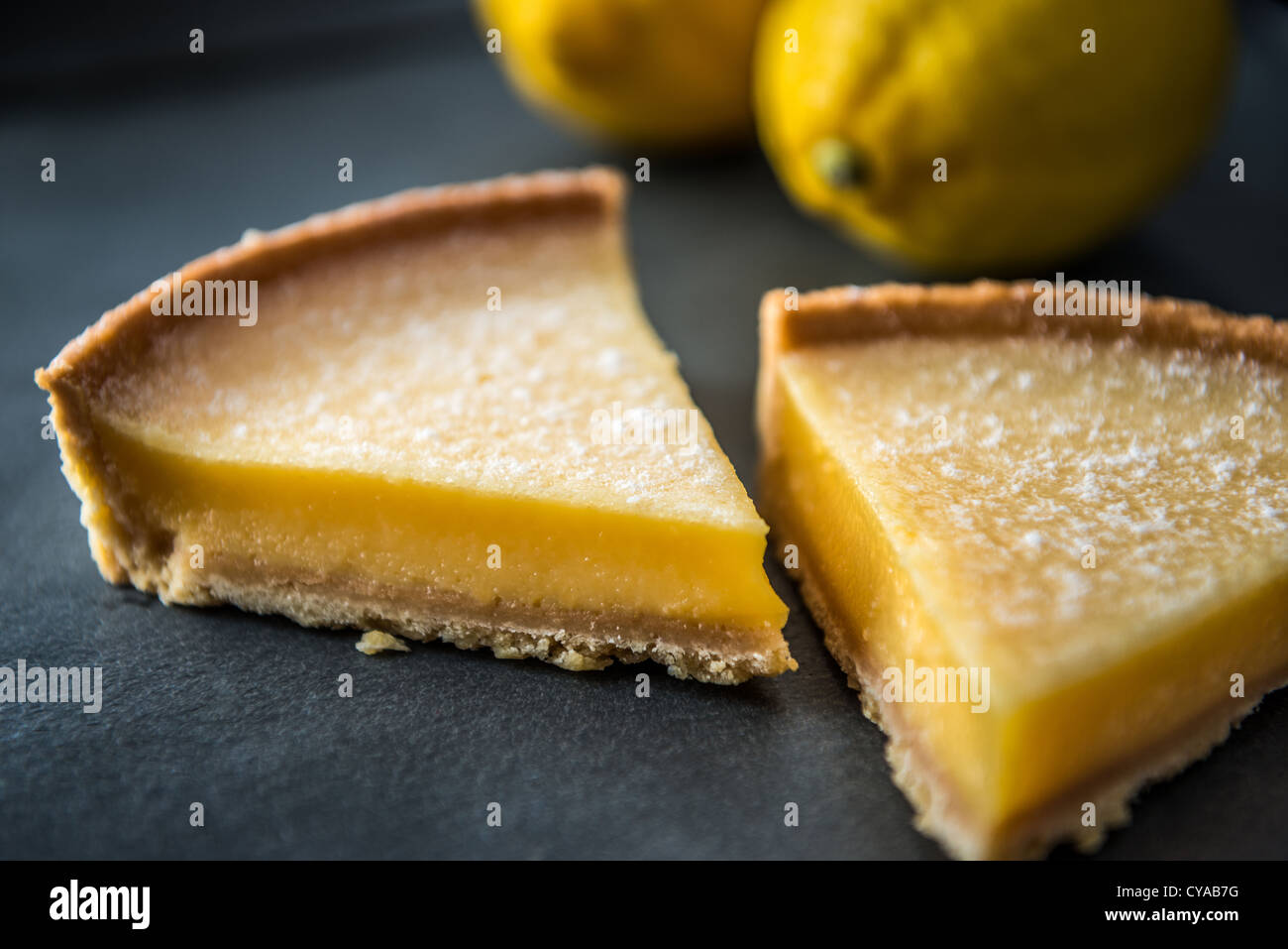 Lemon tart dessert with biscuit base on slate surface with lemons in background. Stock Photo