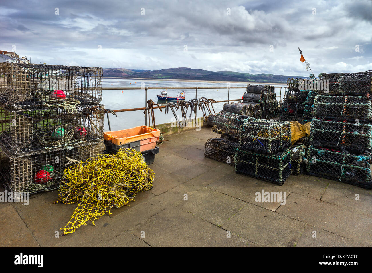 On the quayside is fishing gear comprised of crab and lobster pots, floats, ropes, nets , an inshore fishing boat lies at anchor Stock Photo