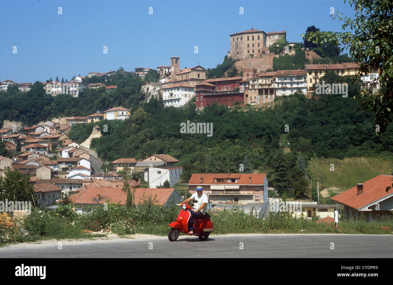 A RIDER ON A RED VESPA PASSES THE VILLAGE OF CASTEL DON ALDO LANGE ITALY Stock Photo