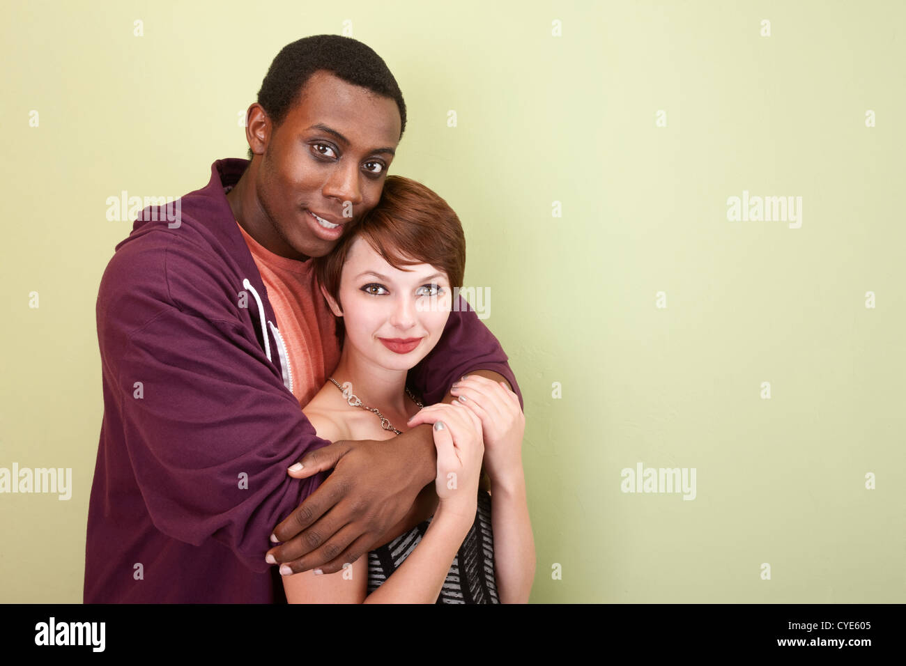 Young mixed race couple hug lovingly in front of a green wall in the studio. Stock Photo